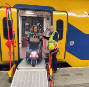 een vrouw die uit de NS trein komt die op een scootmobiel zit. De man met een geel jasje is van de NS reisassistentie help de mevrouw uit de trein te komen.